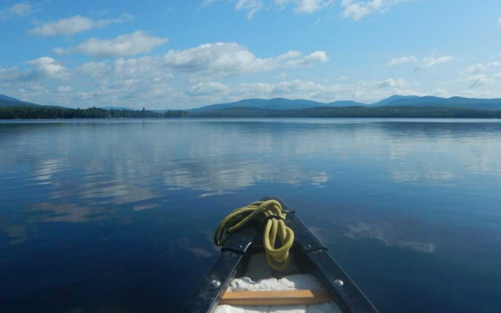 The tip of a canoe appears in the foreground, resting on calm still water that reflects the blue sky dotted with clouds. In the distances, there is a mountainous landscape.